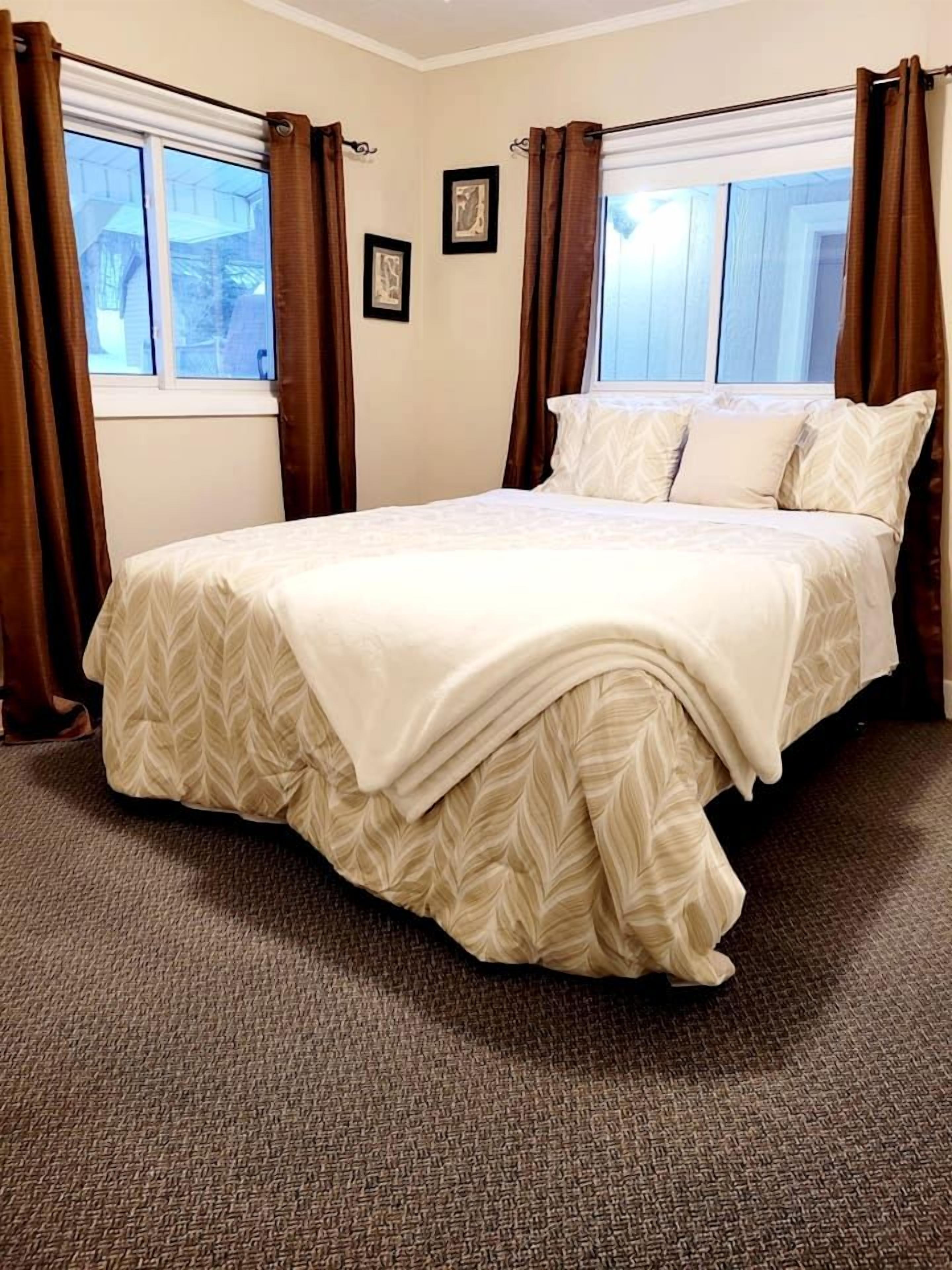 A neatly made bed with a leaf-patterned bedspread, a window with brown curtains, and a carpeted floor.