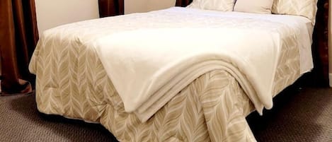 A neatly made bed with a leaf-patterned bedspread, a window with brown curtains, and a carpeted floor.