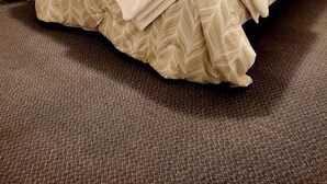 Siististi pedattu sänky, jossa on lehtikuviollinen päiväpeite, ruskeilla verhoilla varustettu ikkuna ja matolla peitetty lattia.