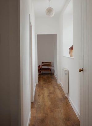 A narrow hallway with wooden flooring, a wooden chair, a radiator, and a door with a knob.