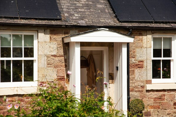 A stone house with a white door and windows, solar panels on the roof, and a garden in the foreground.