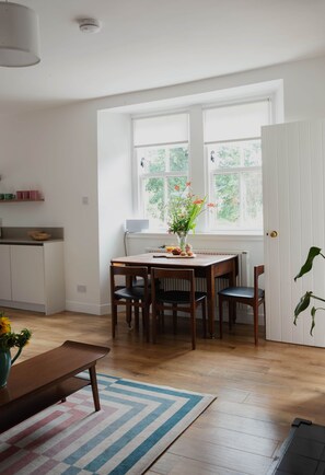 A dining area with a table set for four, a vase of flowers, and a view of greenery through the window.