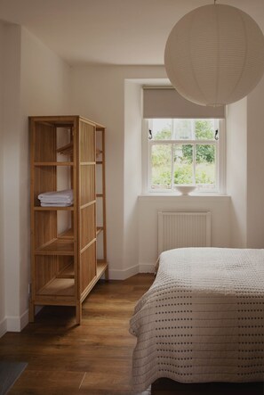 Een slaapkamer met een houten garderobe, een radiator, een raam met uitzicht op groen, en een wit bed met een beddeken met dessin.