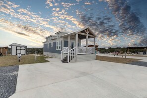 A two-story building with a covered porch and a small deck, surrounded by a concrete patio and grassy areas. - Elegant Tiny Home Retreat with Resort Amenities Near Fredericksburg, TX (Fredericksburg)
