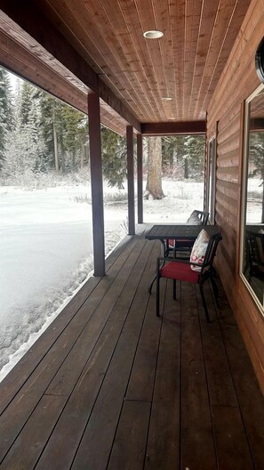 A covered wooden deck with a table and chairs, overlooking a snowy landscape. - Charming Cabin Escape with Hot Tub & Wraparound Porch in McCall, Idaho (McCall)