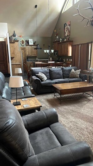 A spacious living room with a leather sofa, a wooden coffee table, and a dining area with a kitchen in the background. - Charming Cabin Escape with Hot Tub & Wraparound Porch in McCall, Idaho (McCall)