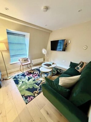 A living room with a green sofa, a coffee table, a floor lamp, a TV mounted on the wall, and a window with blinds. - Stylish One-Bed Apartment in Central Bath Near Train Station & Top Attractions (Bath)