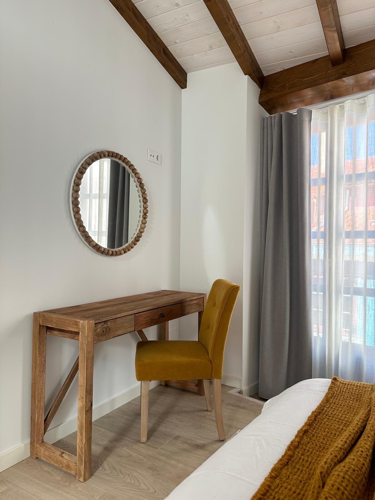 Un dormitori amb escriptori de fusta, una cadira, un llit, una finestra amb cortines i un mirall a la paret.