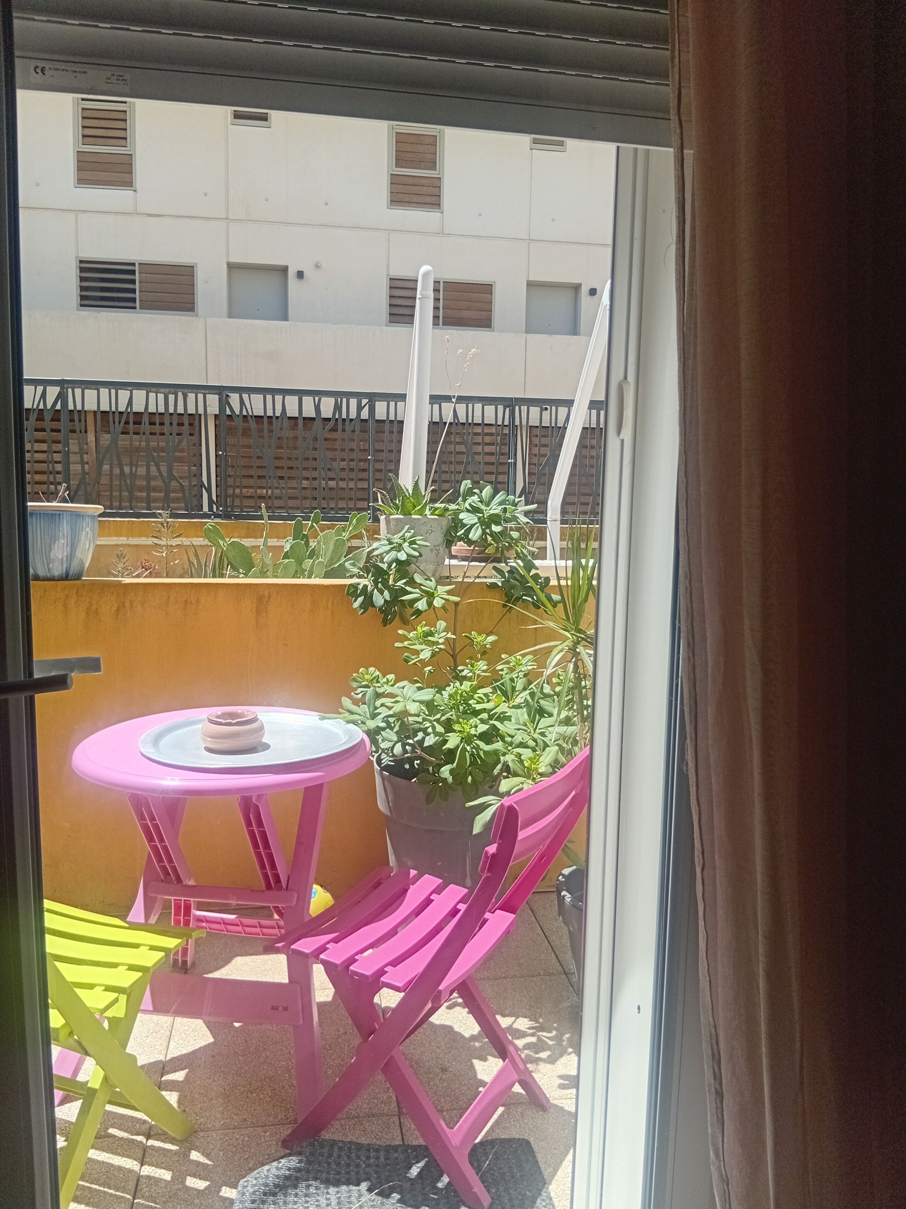 Un balcone con tavolo e sedie, un vaso con una pianta e vista su un edificio sullo sfondo.