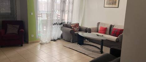 A living room with a sofa, armchair, and coffee table, a window with sheer curtains, and a framed picture on the wall.