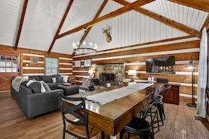 A log cabin interior with a dining area, sofa, fireplace, and a mounted deer head.