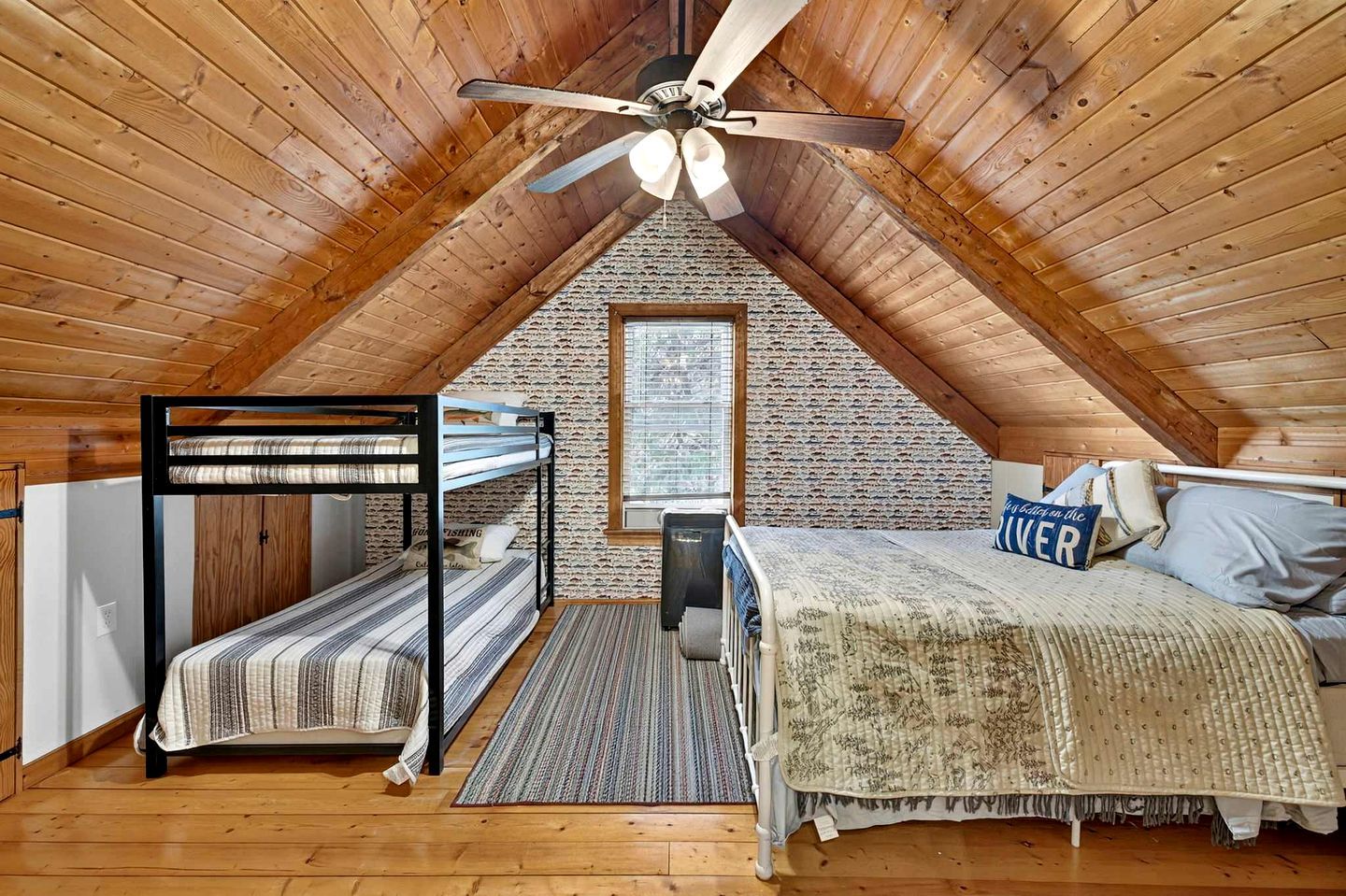 Um quarto de sótão acolhedor com teto inclinado, uma cama beliche, uma cama de solteiro, um ventilador de teto e uma parede de tijolos.