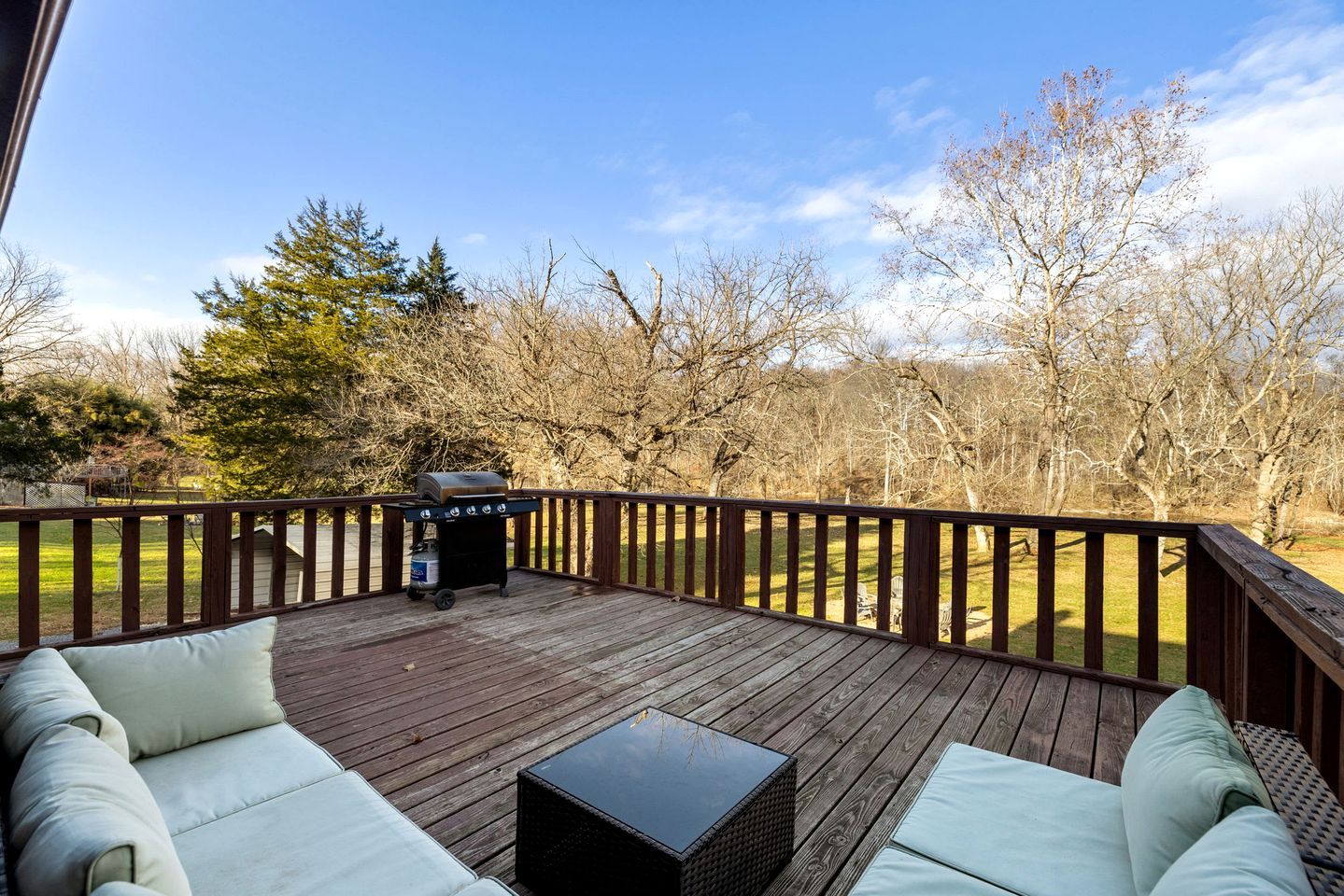 A deck with a grill, seating area, and a view of a grassy field with trees.