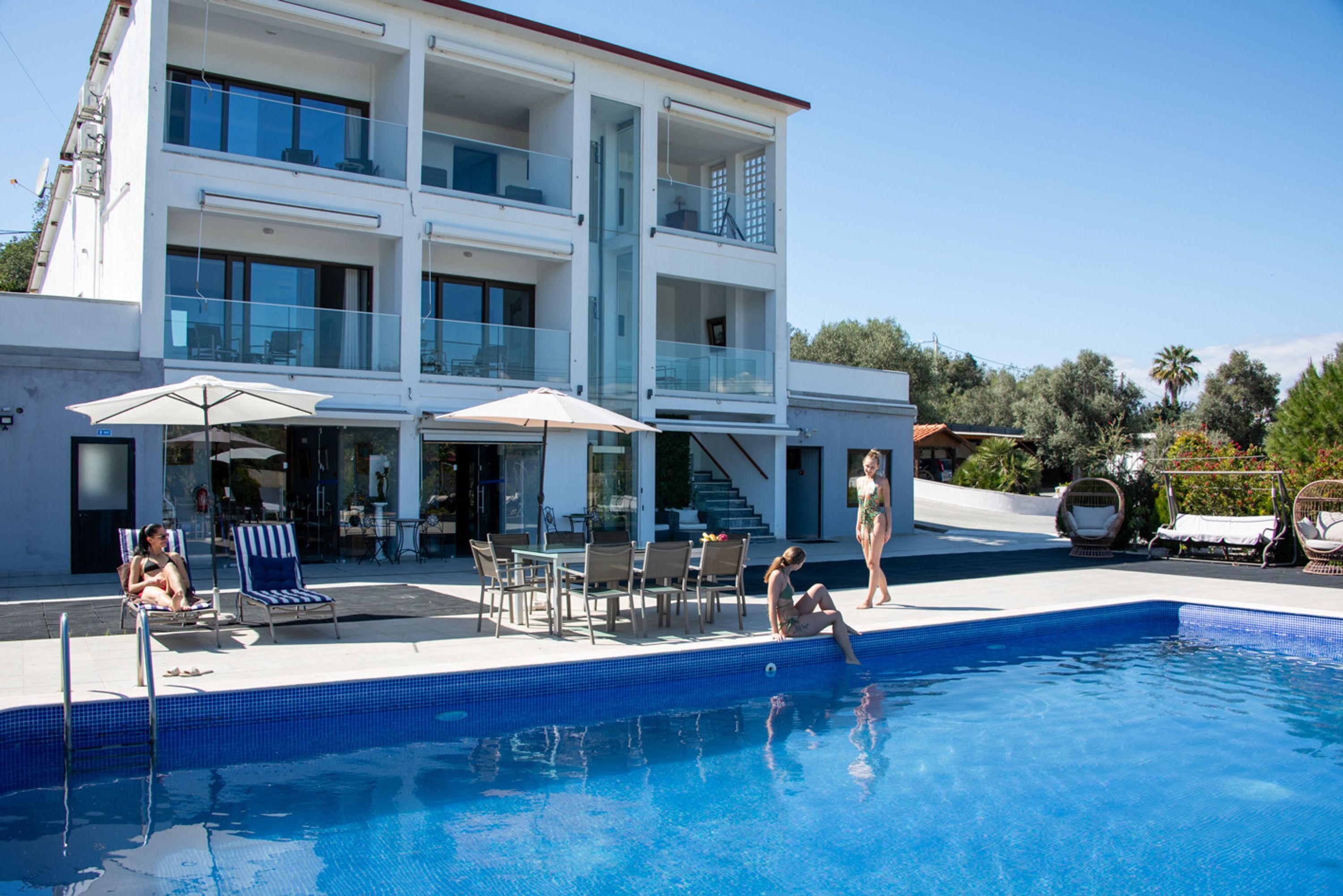A two-story building with a swimming pool, outdoor seating, and a view of the surrounding greenery.