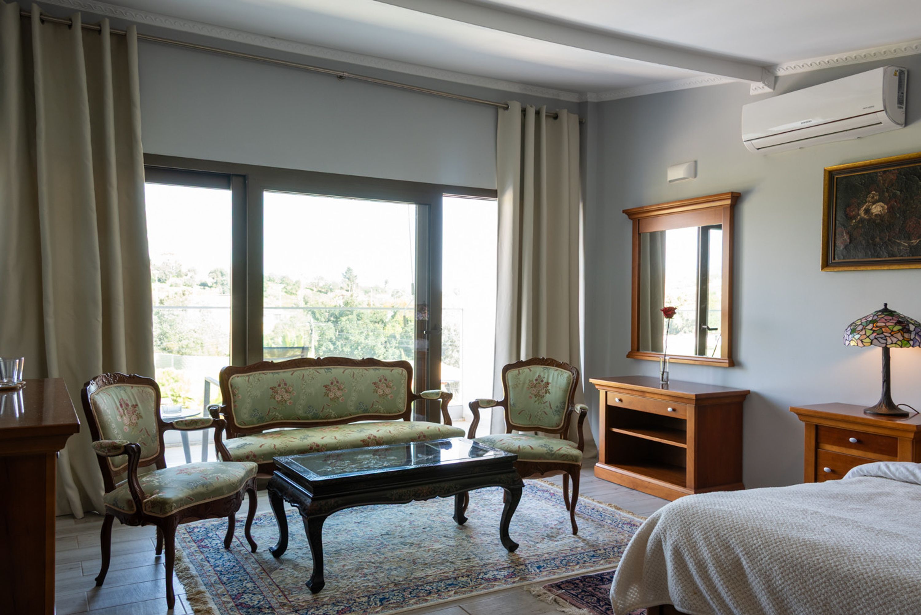 A room with a piano, two armchairs, a wooden cabinet, a bed, and a large window with curtains.