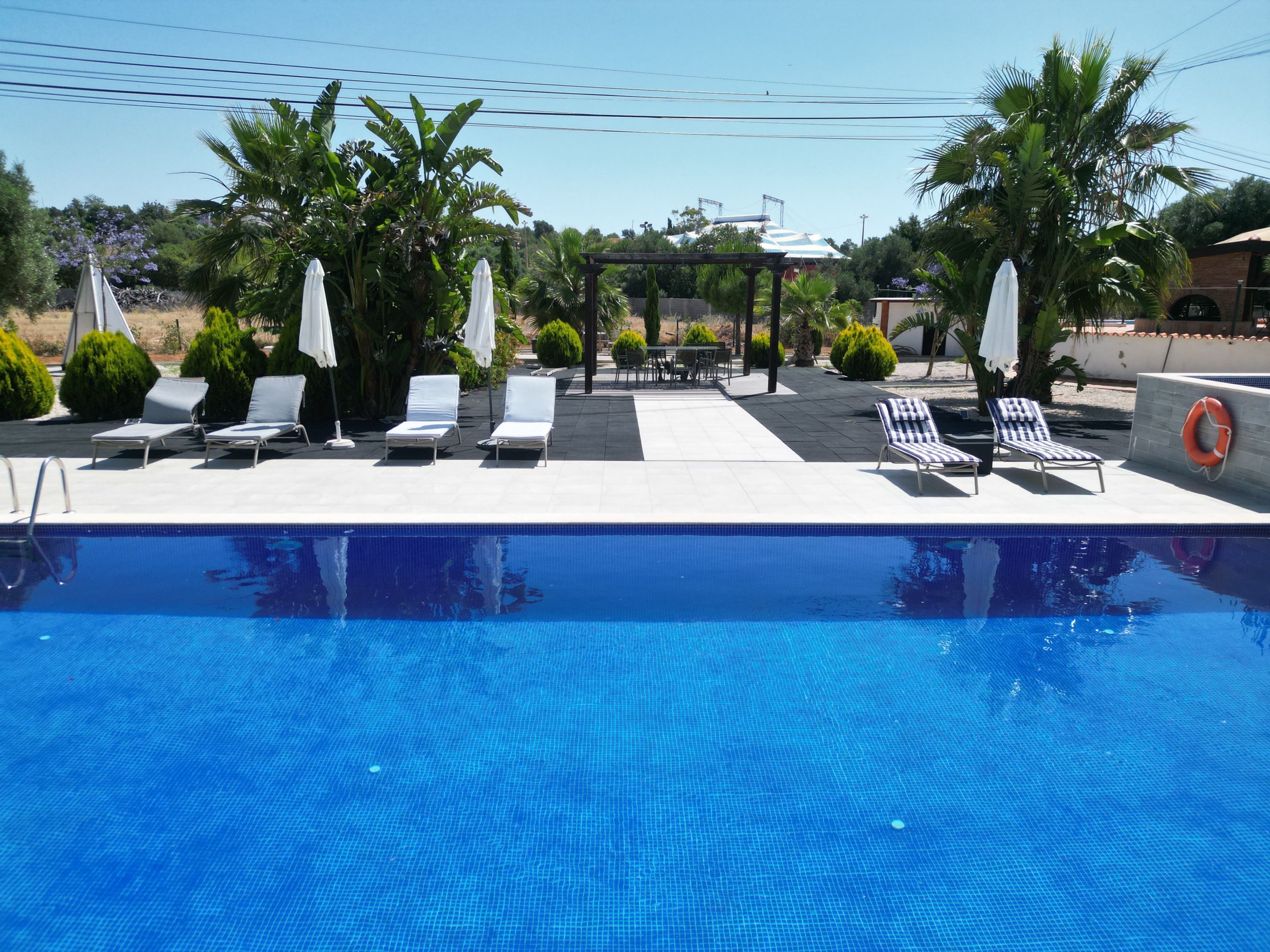 A swimming pool with lounge chairs and umbrellas, surrounded by greenery and a clear sky.