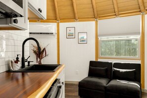 Eine kompakte Küche mit Spüle, integriertem Backofen und Fenster mit Jalousien. An der Wand hängen zwei gerahmte Bilder und es gibt ein dunkelgraues Sofa im Wohnbereich.