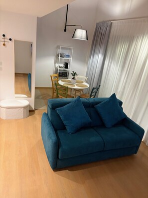 Ein modernes Wohnzimmer mit einem türkisfarbenen Sofa, einem Esstisch mit Stühlen, einem weißen Schrank und einem großen Fenster mit Vorhängen.