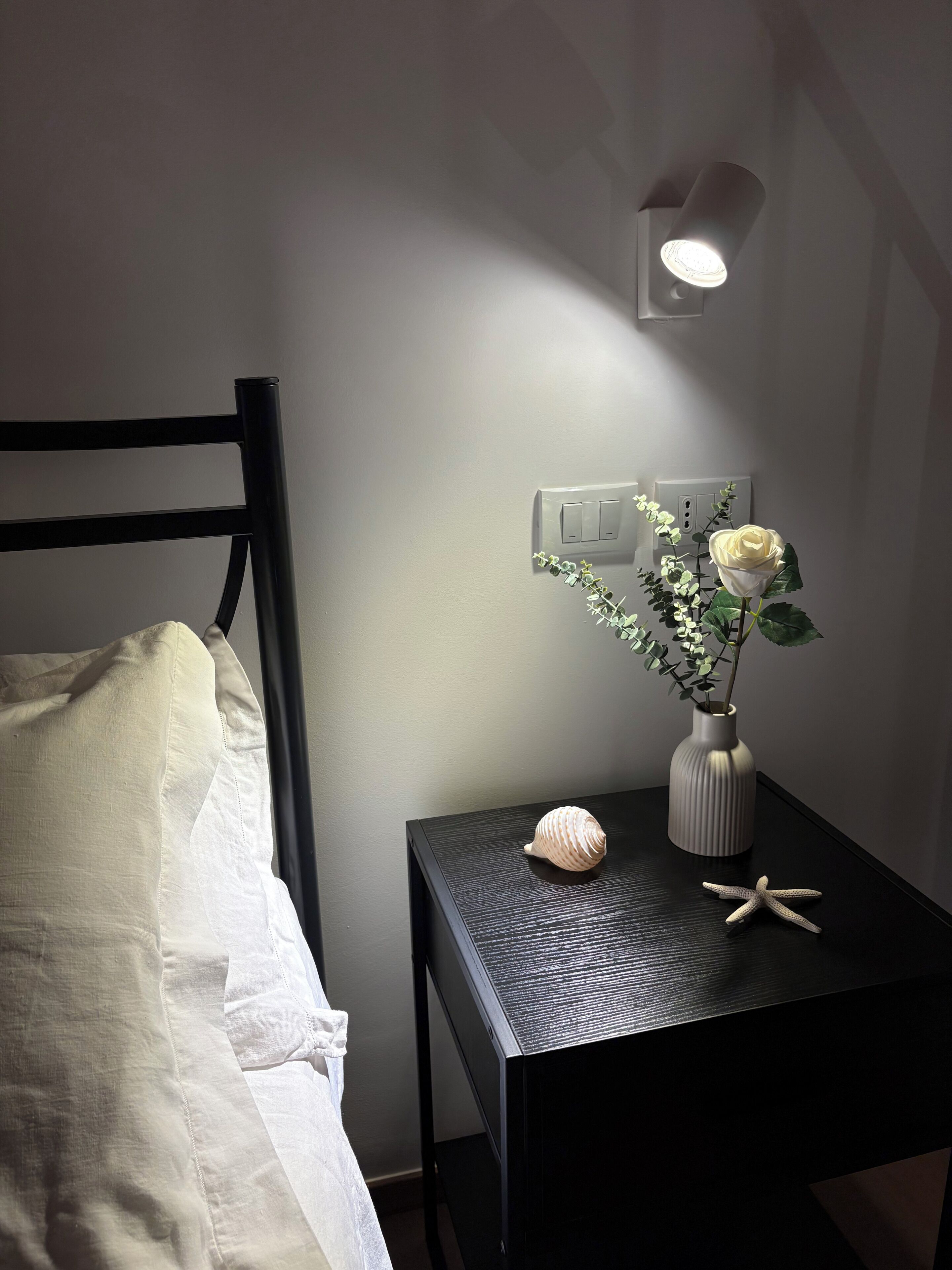 Uma cama com edredom branco, uma mesa de cabeceira com um vaso de flores, uma concha e uma estrela-do-mar.