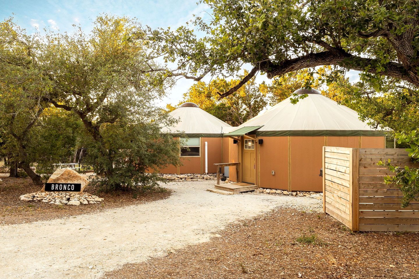 A yurt-style structure with a dirt ground, surrounded by trees and a wooden fence.