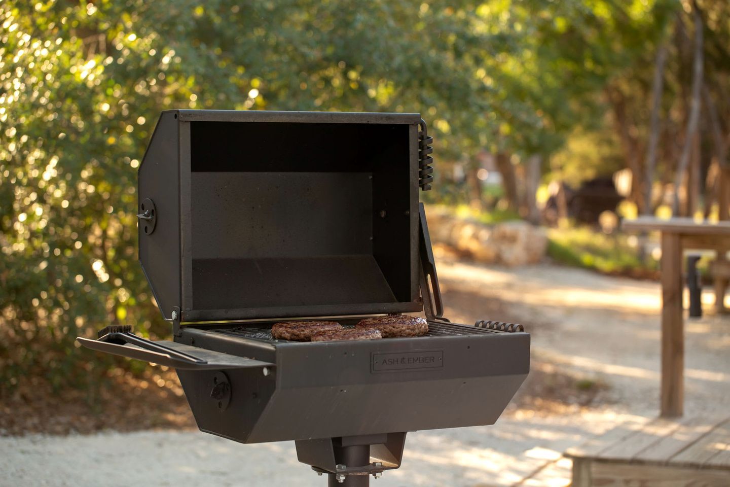 A portable grill with a flat cooking surface and a hinged lid, set outdoors with a natural backdrop.
