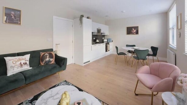 A modern living room with a sofa, a dining table with chairs, a kitchen area, and a large window. - Stylish One-Bed Apartment in the Heart of Historic Bath (Bath)