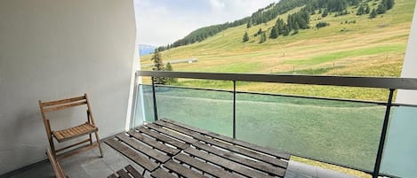 Un balcón con banca de madera y una silla, con vista a un paisaje verde con árboles y una colina.