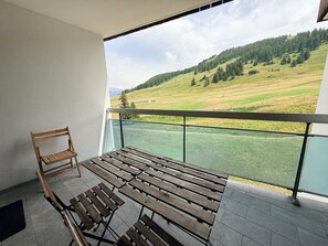 Ein Balkon mit einer hölzernen Bank und einem Stuhl, mit Blick auf eine grüne Landschaft mit Bäumen und einer Hügelkette.