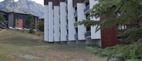 Un bâtiment moderne doté de plusieurs balcons, entouré d’une zone verdoyante et d’arbres, avec des montagnes en arrière-plan.