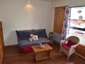 A cozy living room with a grey sofa, a wicker chair, a small wooden table, and a window with a view of houses. - Studio near slopes - 4 guests (Montgenèvre)