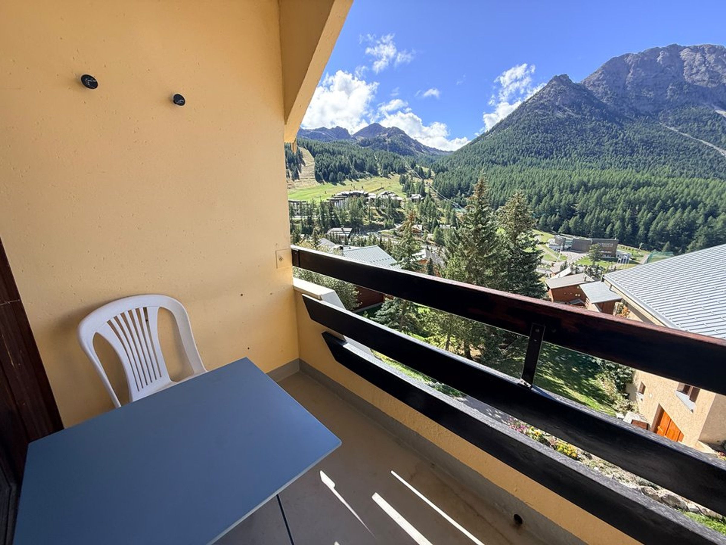 Un balcon avec une table et des chaises, donnant sur une vallée bordée de montagnes et comprenant un petit village.