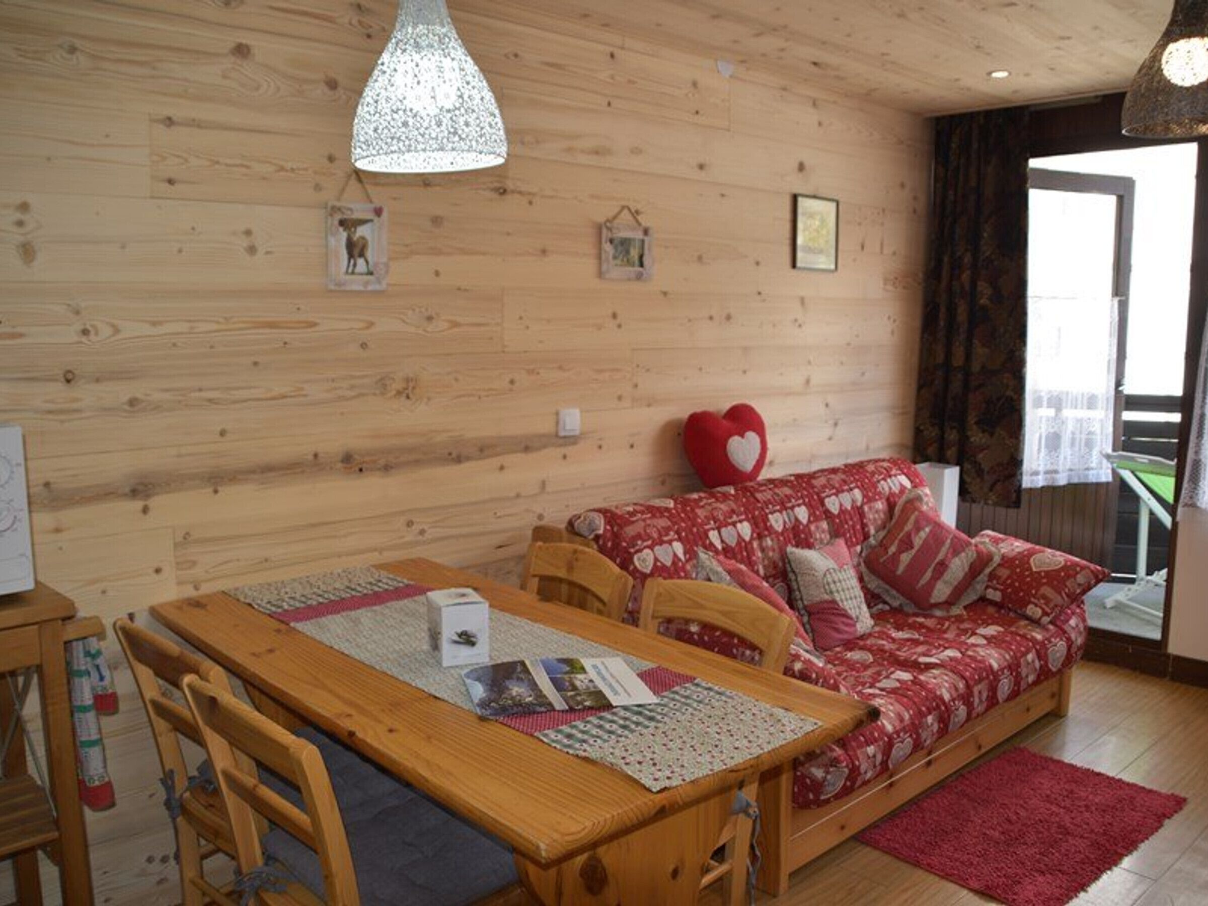 A cozy room with a wooden floor, a wooden dining table with chairs, a red sofa with heart-shaped pillows, and a hanging light fixture.