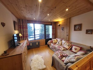Ein gemütliches Zimmer mit Holzbalkendecke, einer Couch mit gemusterten Kissen, einem kleinen Teppich und einem Fenster mit Vorhängen.