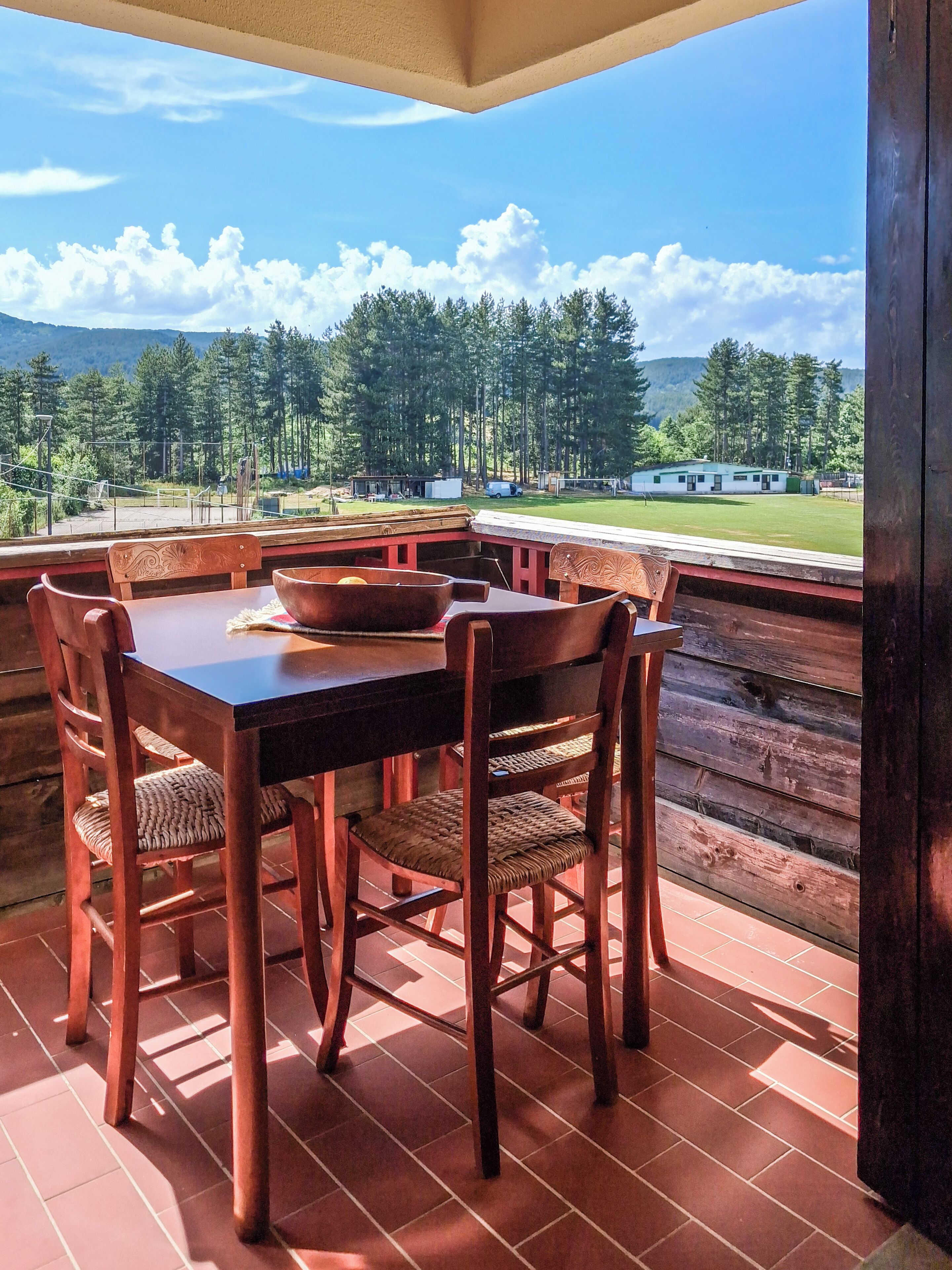 A wooden table with chairs on a balcony with a view of a green field and trees.