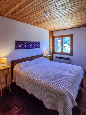 A bedroom with a wooden ceiling, a bed with a white textured blanket, a nightstand with a lamp, a decorative wall hanging, and a window with a view. - Dolce Casa - Residence La Montanara (Camigliatello Silano)