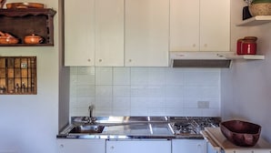 A kitchen with white cabinets, a stainless steel sink, and a stove. There are shelves with kitchenware and a decorative vase. - Dolce Casa - Residence La Montanara (Camigliatello Silano)