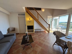 A living room with a sofa, a coffee table with wine glasses, a fireplace, a staircase, and a balcony with a table and chairs. - Apartment sea view - 6 guests (Saint-Cyr-sur-Mer)