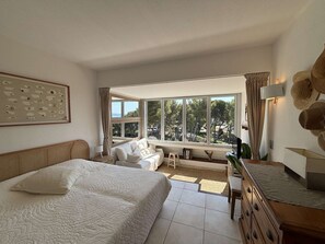 Ein Schlafzimmer mit einem großen Bett, einer Couch, einem Schreibtisch und Blick auf Bäume durch ein großes Fenster.