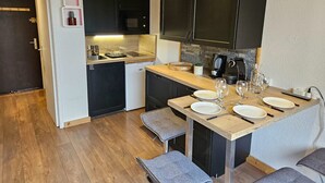 Kompakti keittiö, jossa on tummat kaapit, mikroaaltouuni ja kahvinkeitin. Siellä on pieni ruokailutila, jossa on pöytä kahdelle hengelle ja penkki.