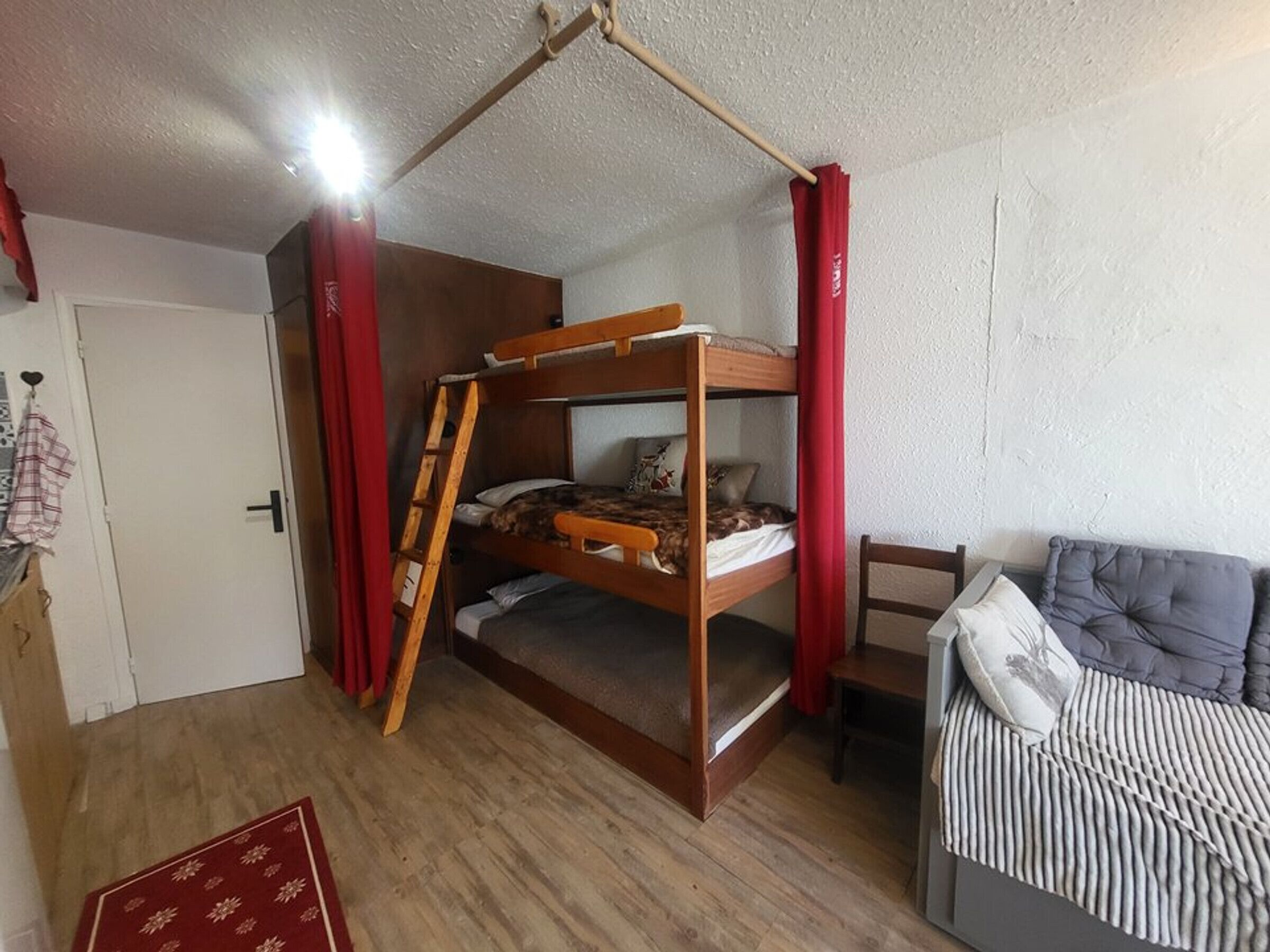 A bunk bed with a ladder, a small table, and a sofa in a room with wooden flooring and white walls.