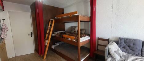 A bunk bed with a ladder, a small table, and a sofa in a room with wooden flooring and white walls.