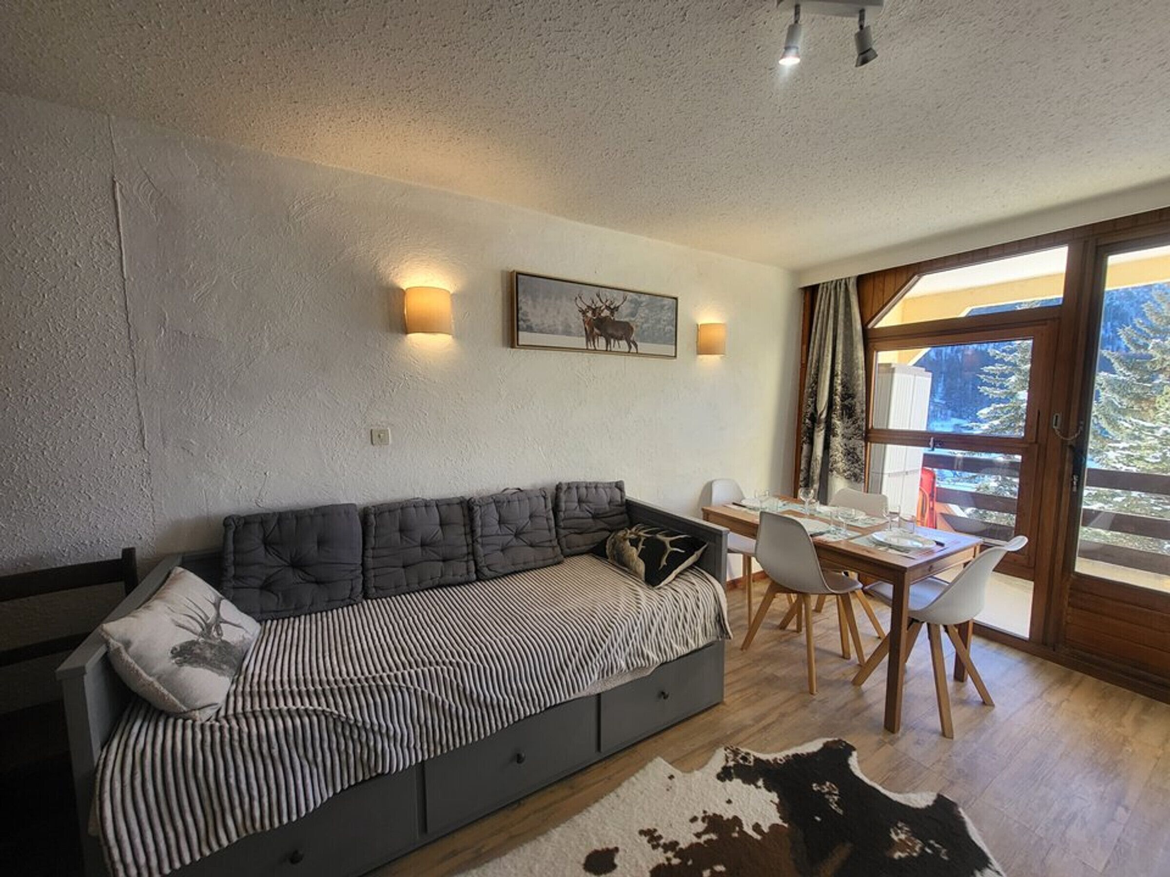 Un salon moderne avec un canapé, une table à manger et des chaises. Une grande fenêtre offre une vue sur les arbres et une montagne.
