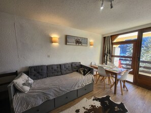 Ein modernes Wohnzimmer mit Sofa, Esstisch und Stühlen. Ein großes Fenster bietet Blick auf Bäume und einen Berg.