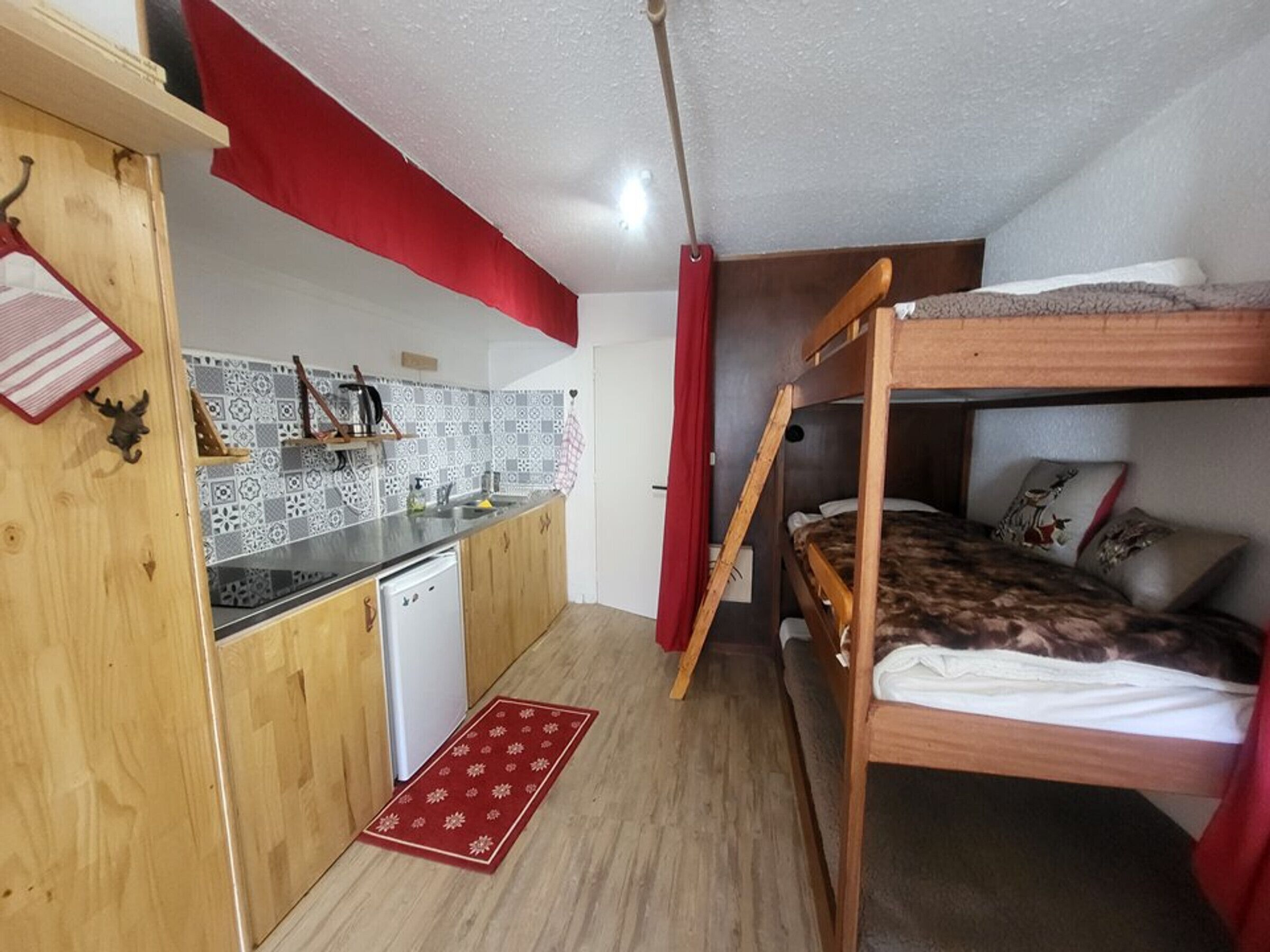 Une petite pièce avec un lit superposé, un coin cuisine et un rideau rouge.