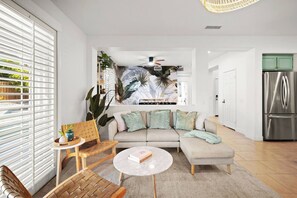 Una sala de estar moderna con sofá, mesa de centro y sillas, una cocina con refrigerador, y un ventanal con persianas.