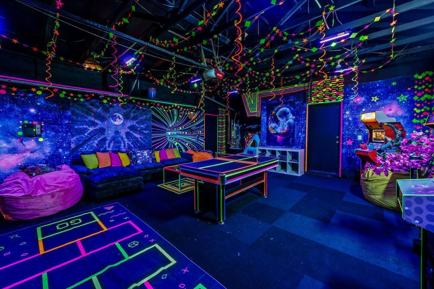 A neon-lit room with a gaming table, bean bag, and a wall with a space-themed mural.