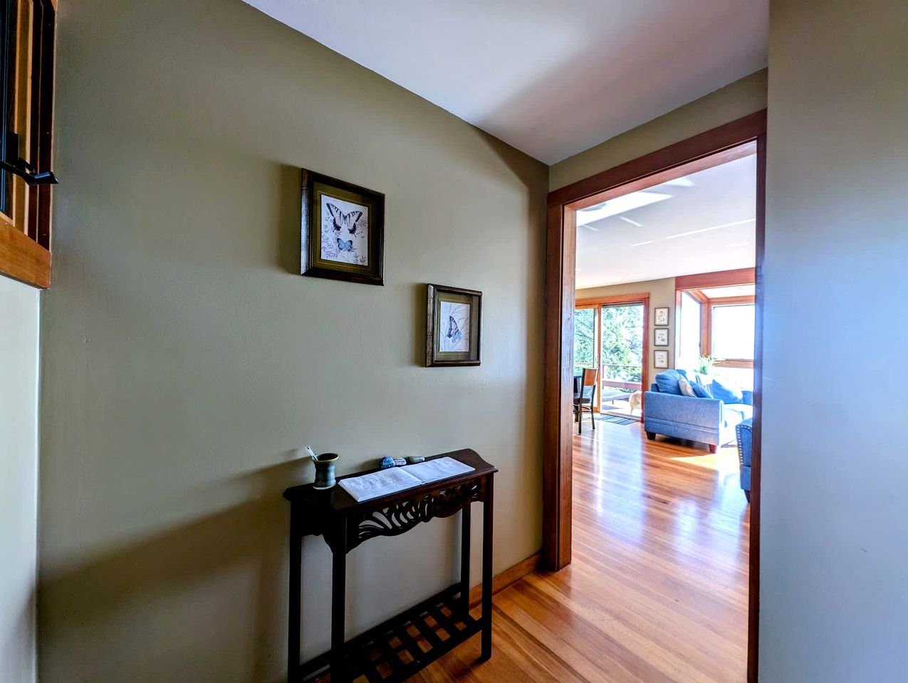 A hallway with a wooden floor, a small table with a vase and papers, and two framed pictures on the wall.