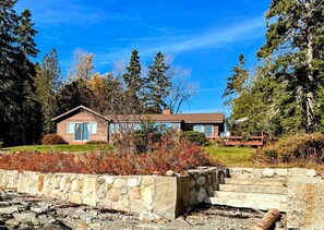 A two-story house with a stone retaining wall in the foreground. - Charming Waterfront Sea Cottage with Private Beach near Bar Harbor, Hancock Maine (Hancock)