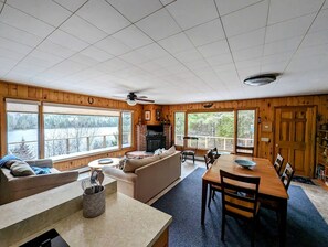 A cabin interior with a fireplace, a dining area, a sofa, and large windows overlooking a natural landscape. - Secluded Oceanfront Cottage with Wrap-Around Deck near Acadia National Park, Sullivan, Maine (Sullivan)