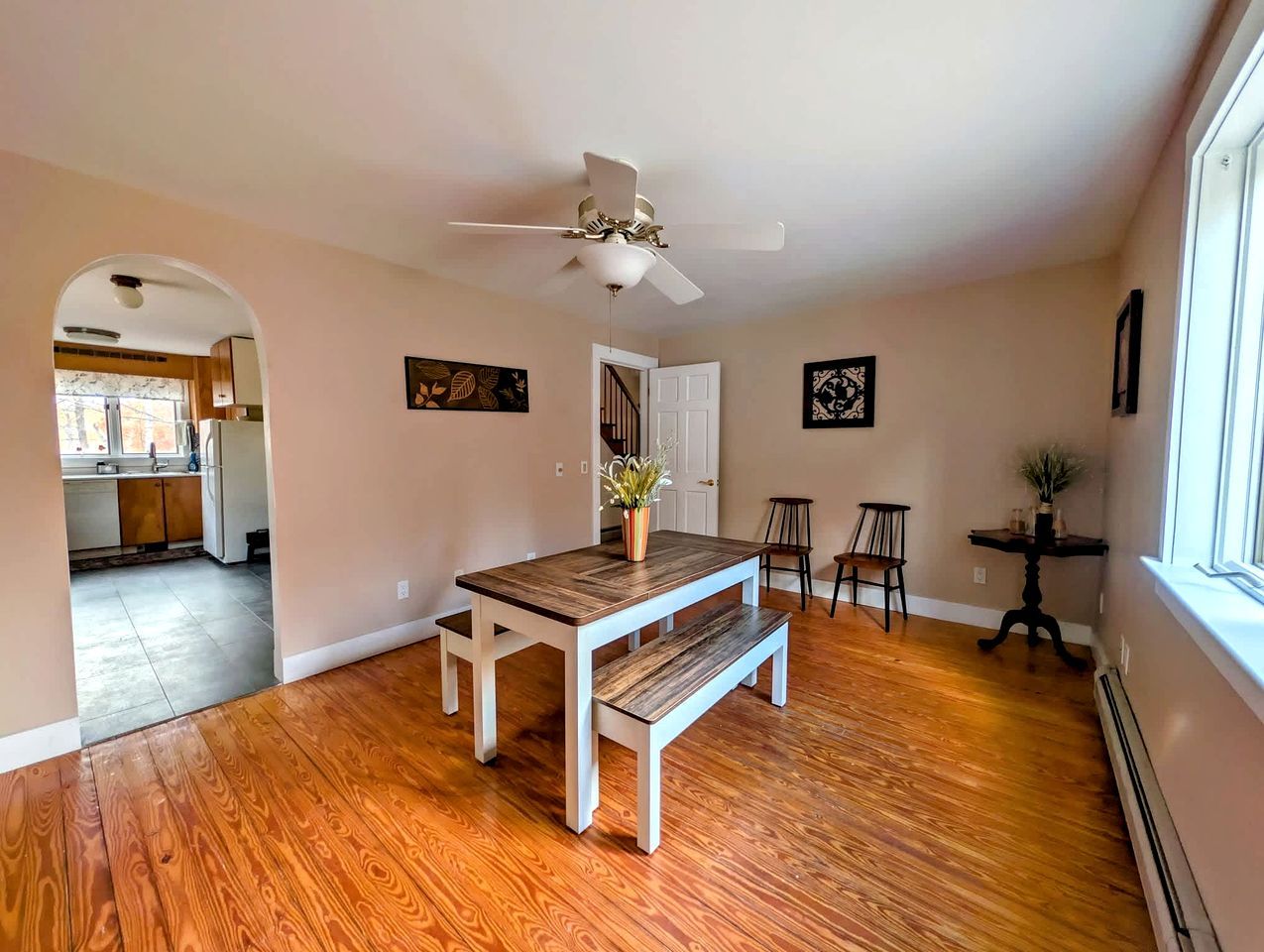 Une pièce spacieuse avec un plancher en bois, une table à manger avec des bancs, un ventilateur de plafond et une vue sur un coin cuisine.